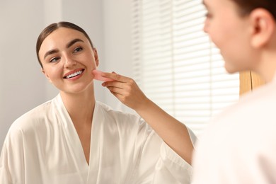 Beautiful young woman doing facial massage with gua sha tool near mirror at home