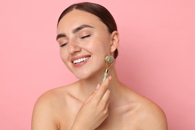 Beautiful young woman doing facial massage with roller on pink background