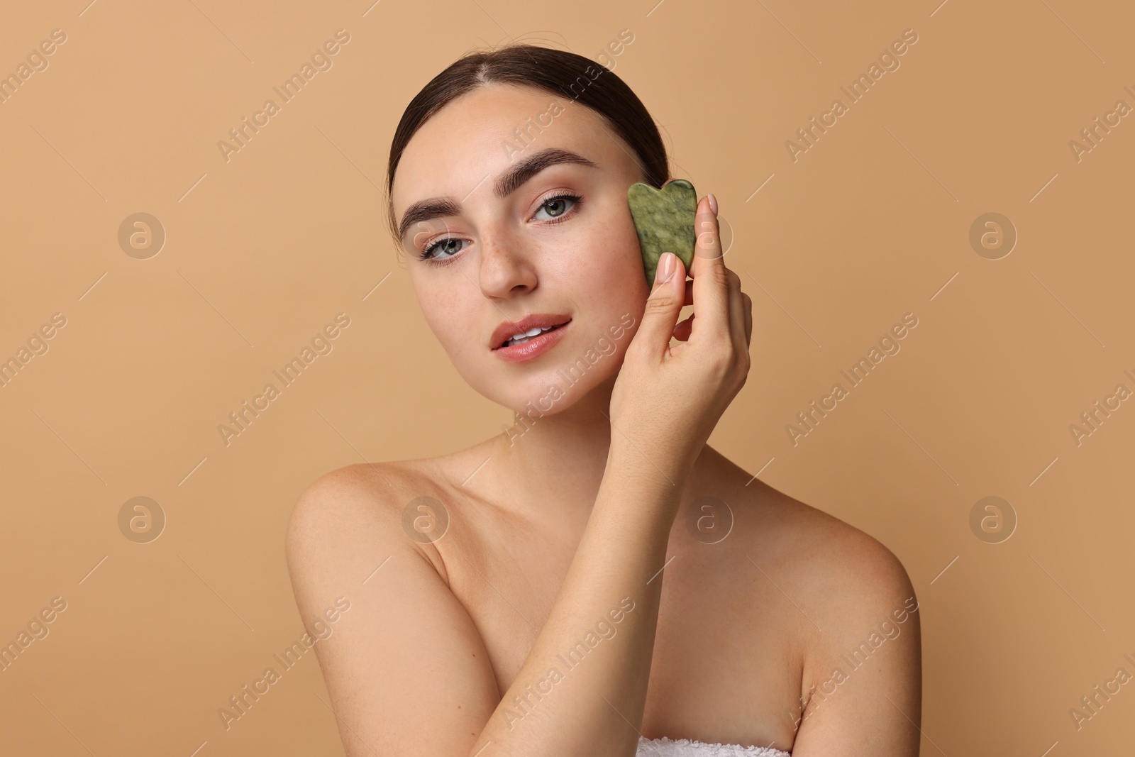 Photo of Beautiful young woman doing facial massage with gua sha tool on beige background