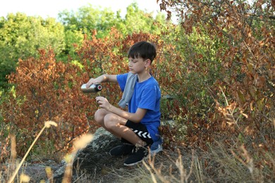 Photo of Little boy pouring tea from thermos into cup among trees outdoors