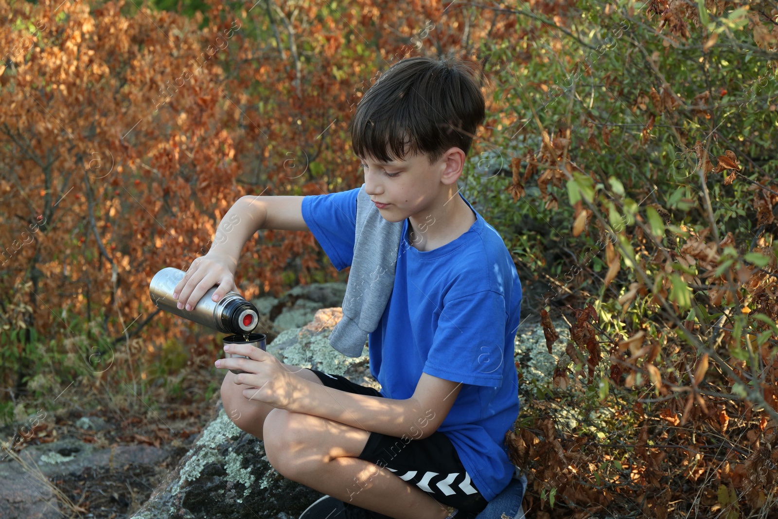 Photo of Little boy pouring tea from thermos into cup among trees outdoors