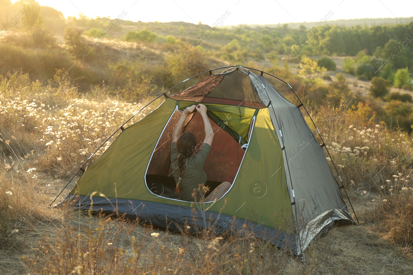 Photo of Woman fixing entrance of camping tent among plants outdoors on sunny day, back view