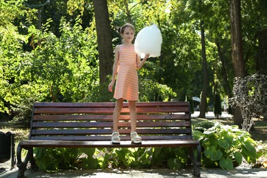 Photo of Little girl with sweet cotton candy on wooden bench in park