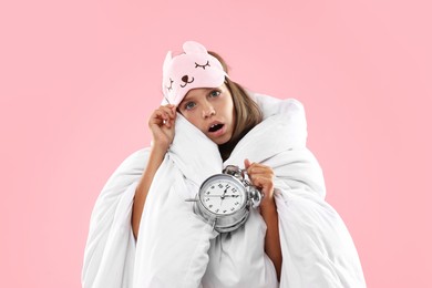 Photo of Overslept girl with sleep mask and alarm clock wrapped in blanket on pink background