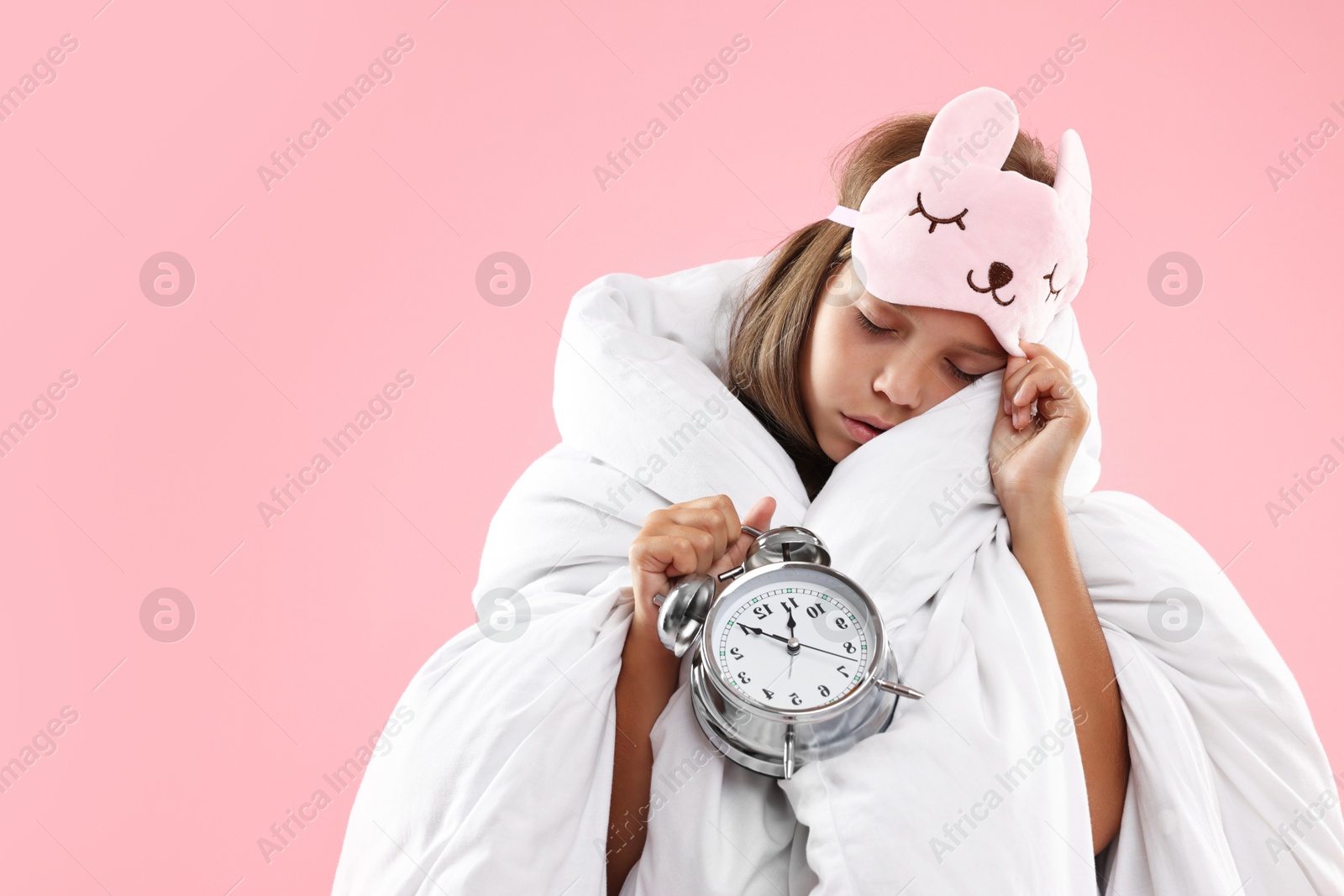 Photo of Overslept girl with sleep mask and alarm clock wrapped in blanket on pink background, space for text