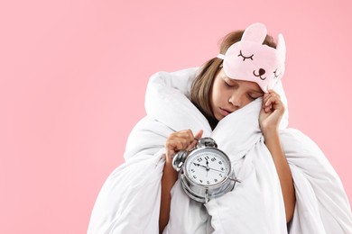 Photo of Overslept girl with sleep mask and alarm clock wrapped in blanket on pink background, space for text