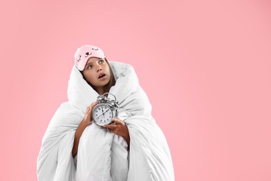 Overslept girl with sleep mask and alarm clock wrapped in blanket on pink background, space for text