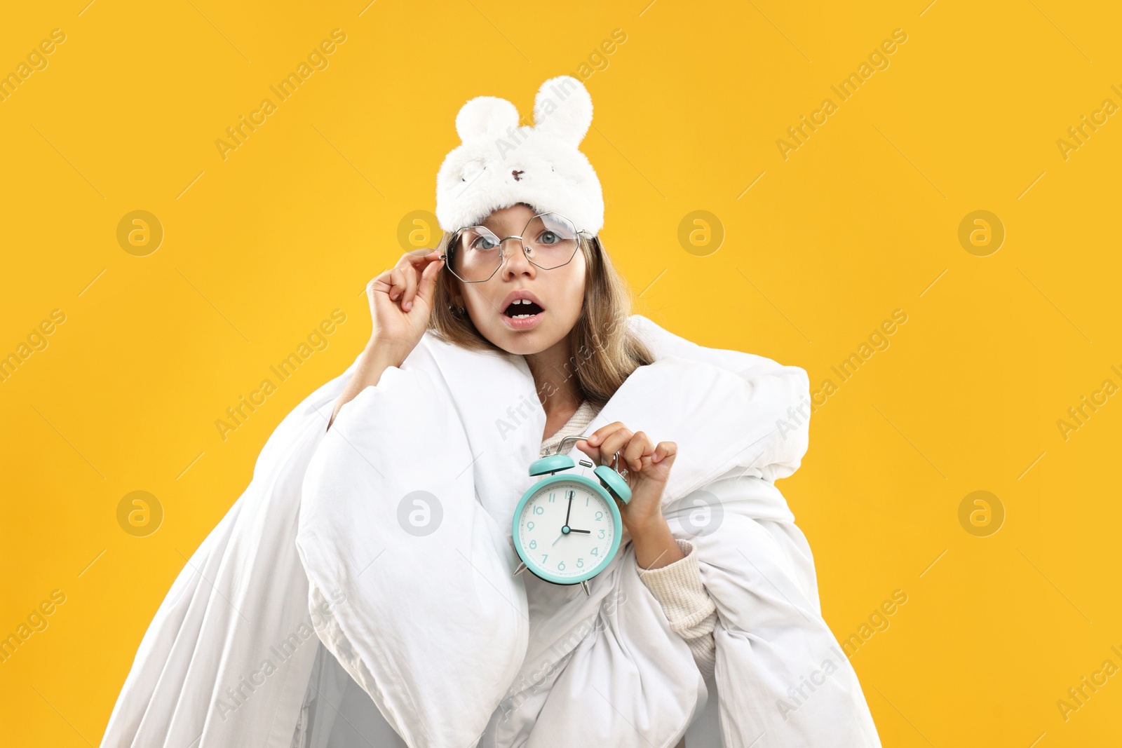 Photo of Overslept girl with sleep mask and alarm clock wrapped in blanket on orange background