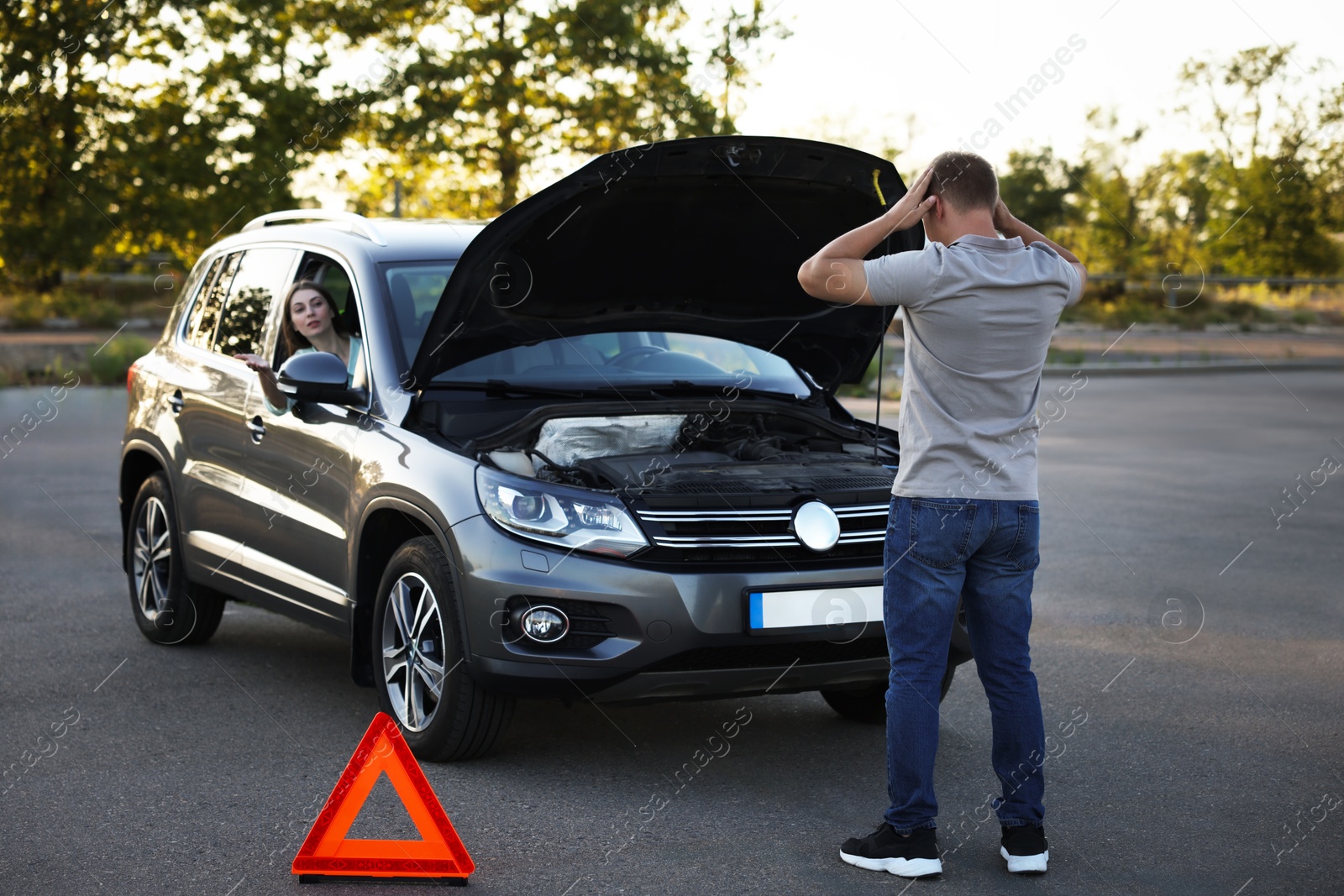 Photo of Stressed man standing near broken car while woman sitting in auto outdoors