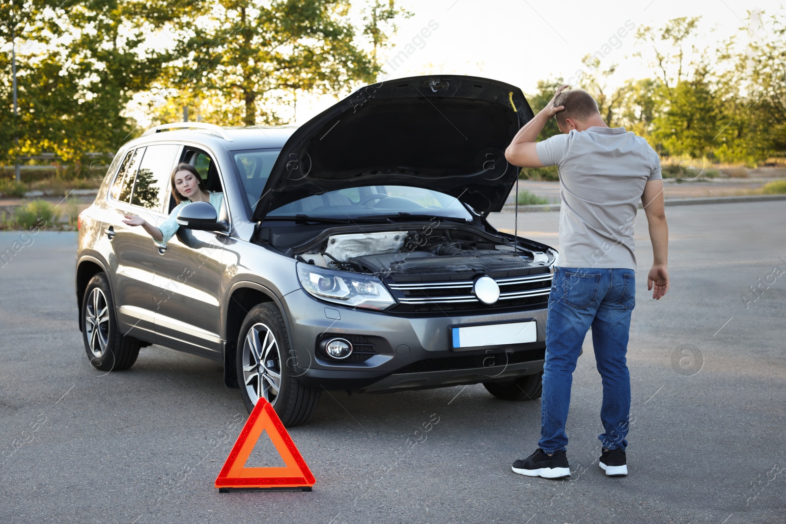 Photo of Stressed man standing near broken car while woman sitting in auto outdoors