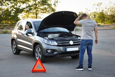 Stressed man standing near broken car while woman sitting in auto outdoors