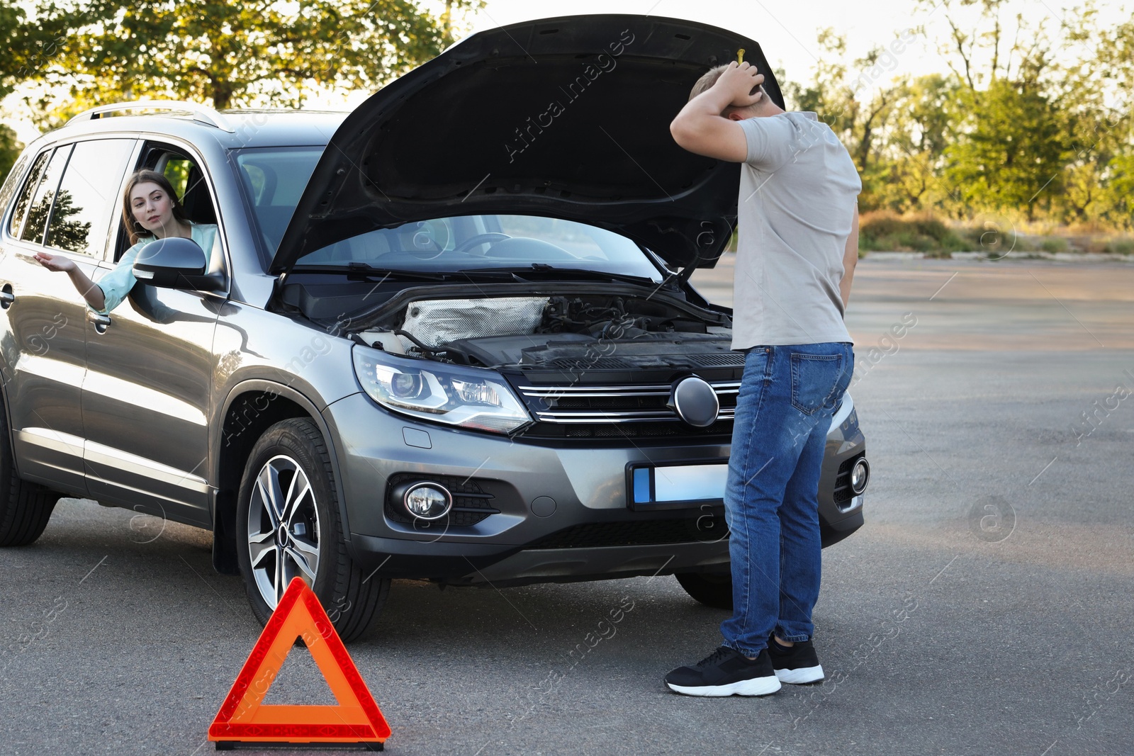 Photo of Stressed man standing near broken car while woman sitting in auto outdoors