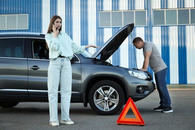 Man examining broken auto while woman calling to car service outdoors