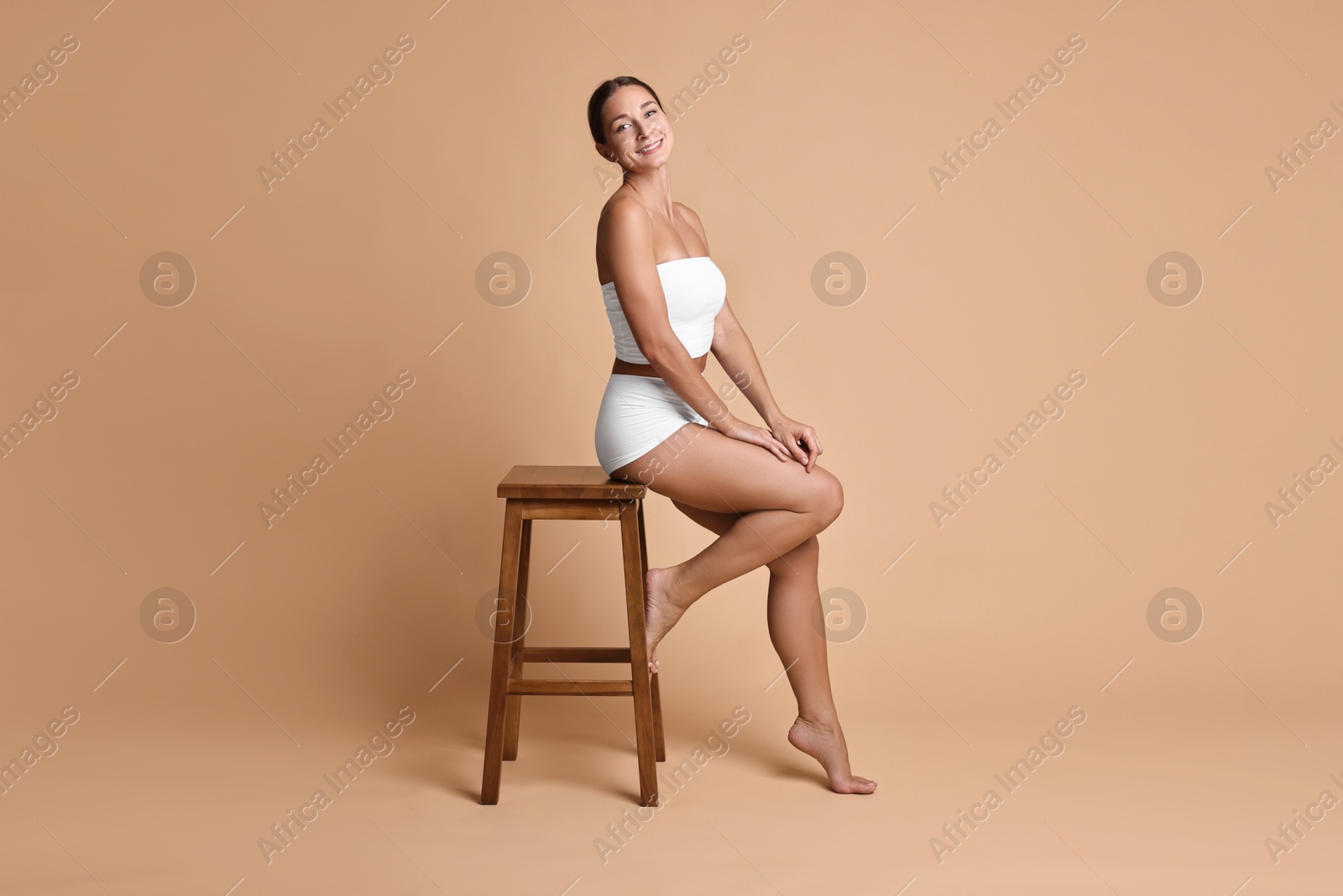 Photo of Smiling woman with perfect skin in underwear posing on stool against beige background. Body care