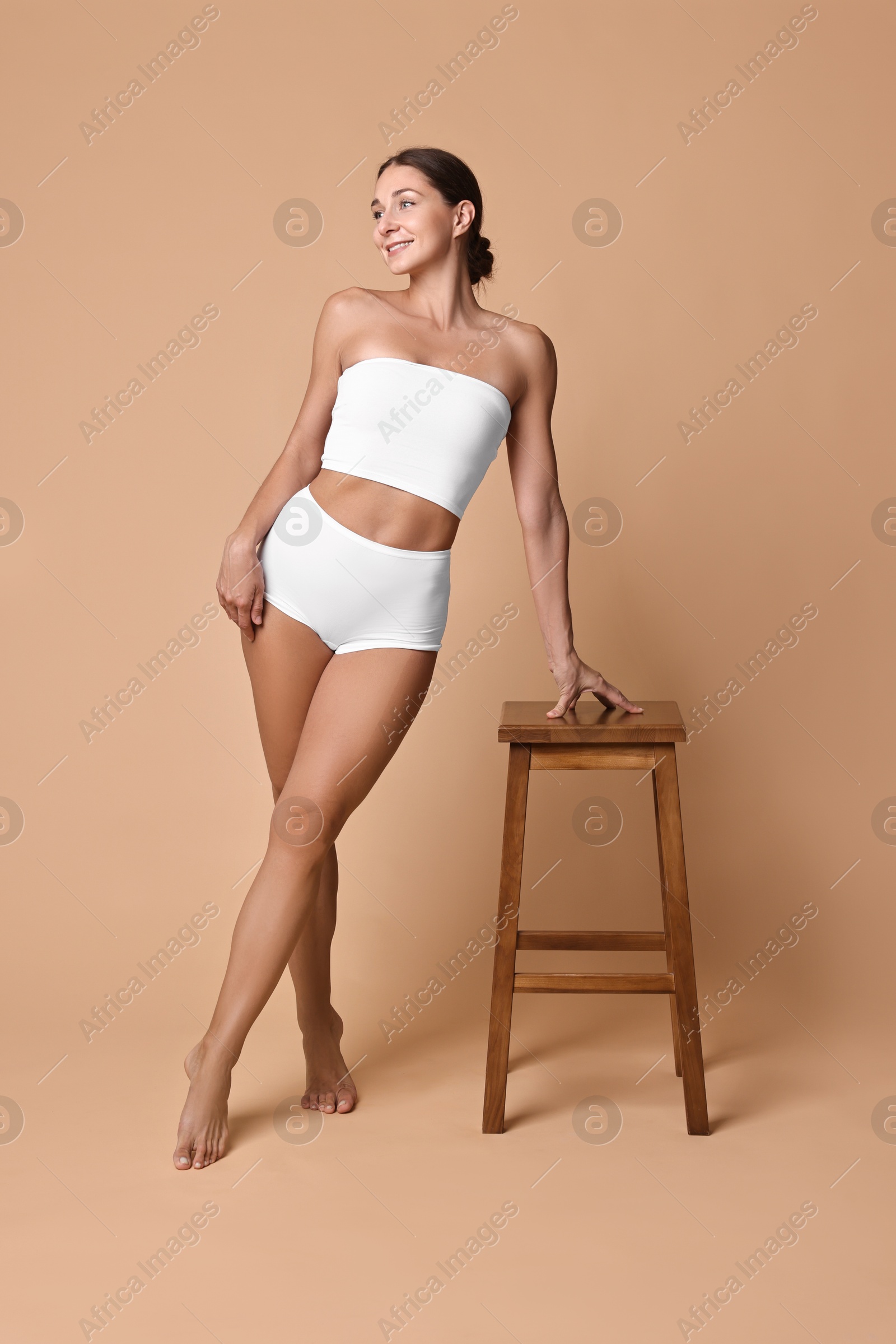 Photo of Smiling woman with perfect skin in underwear posing with stool against beige background. Body care