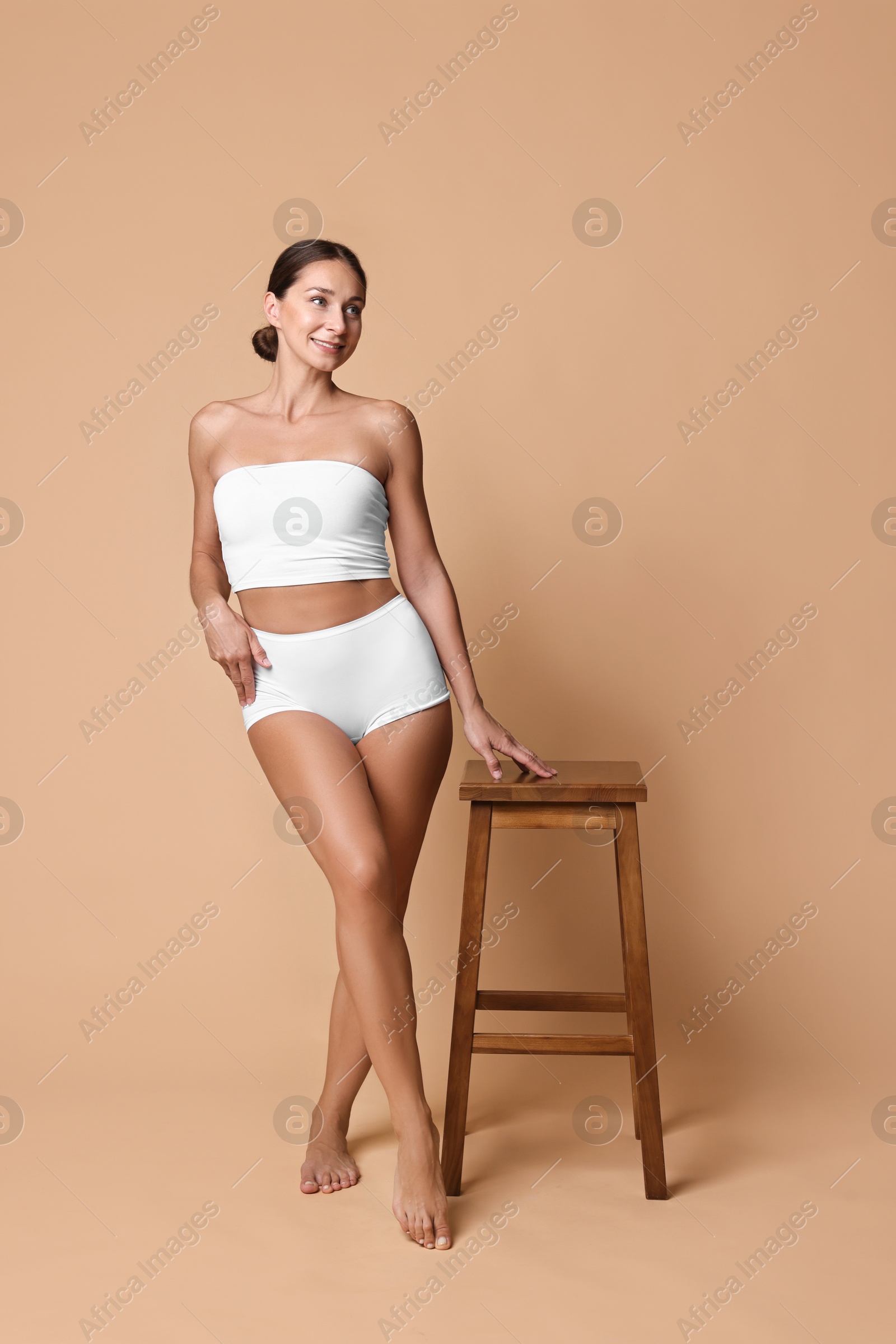 Photo of Smiling woman with perfect skin in underwear posing with stool against beige background. Body care