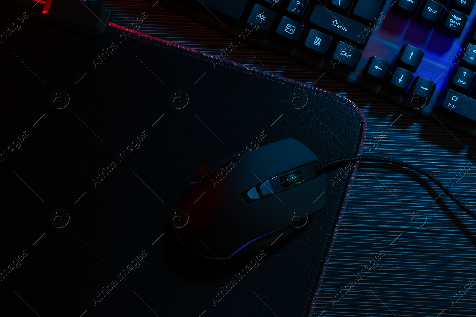 Photo of Computer mouse, mousepad and keyboard on dark wooden table in neon lights, flat lay