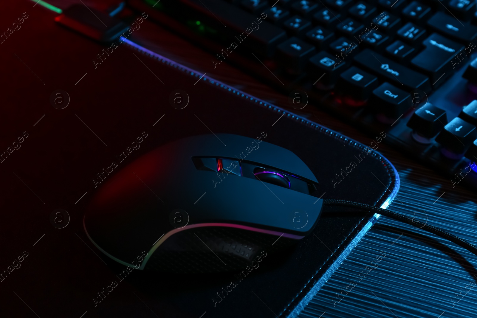 Photo of Computer mouse, mousepad and keyboard on dark wooden table in neon lights, closeup