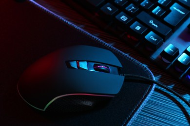 Photo of Computer mouse, mousepad and keyboard on dark wooden table in neon lights, closeup