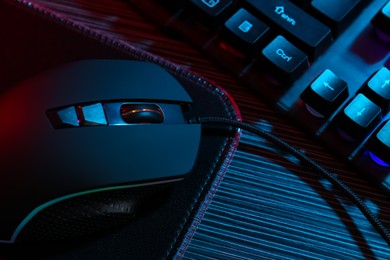 Photo of Computer mouse, mousepad and keyboard on dark wooden table in neon lights, closeup