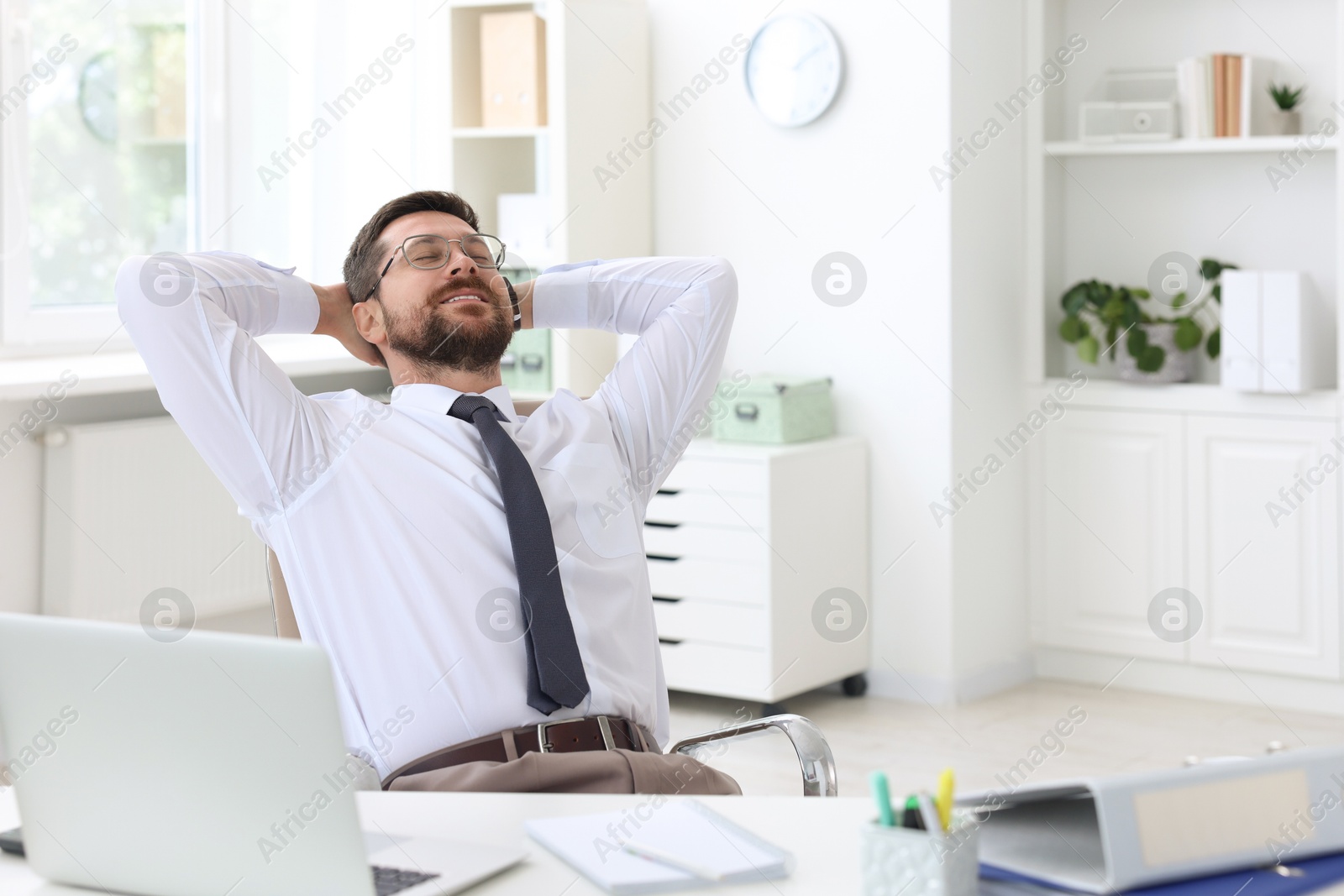 Photo of Smiling businessman sitting at table in office with hands behind his head. Break time