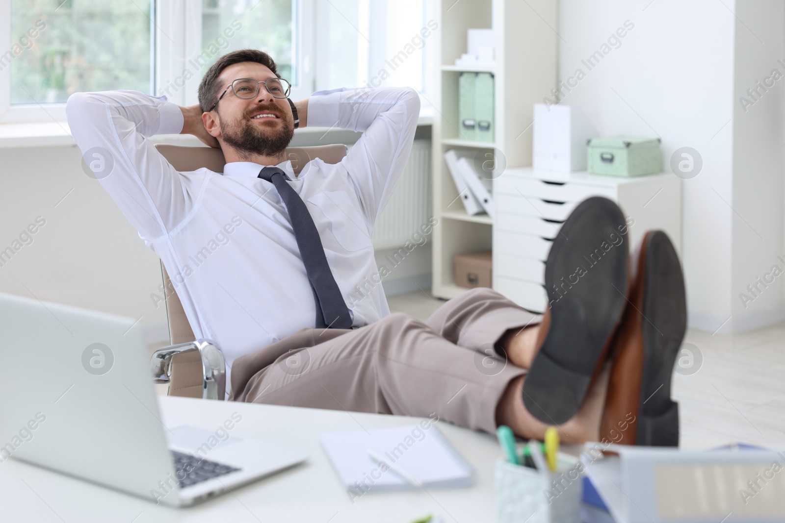 Photo of Smiling businessman with hands behind his head holding legs on table in office. Break time