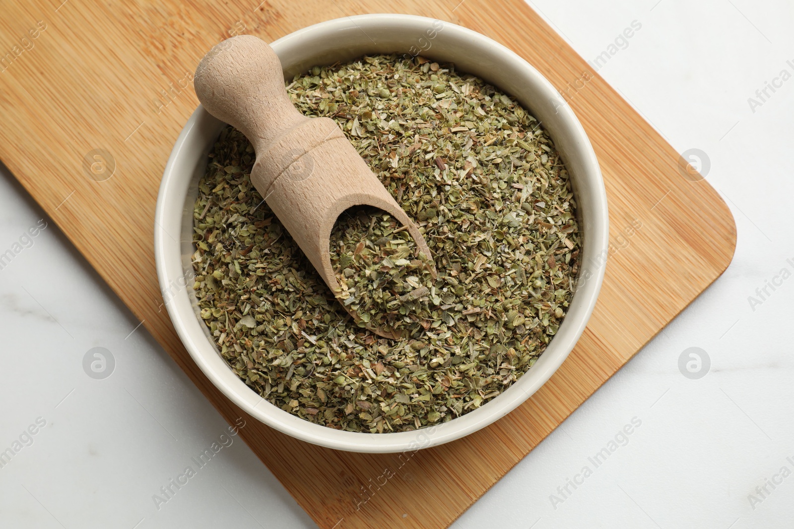 Photo of Dried oregano in bowl and scoop on white marble table, top view