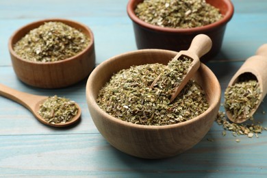 Dried oregano in bowls, scoops and spoon on light blue wooden table, closeup