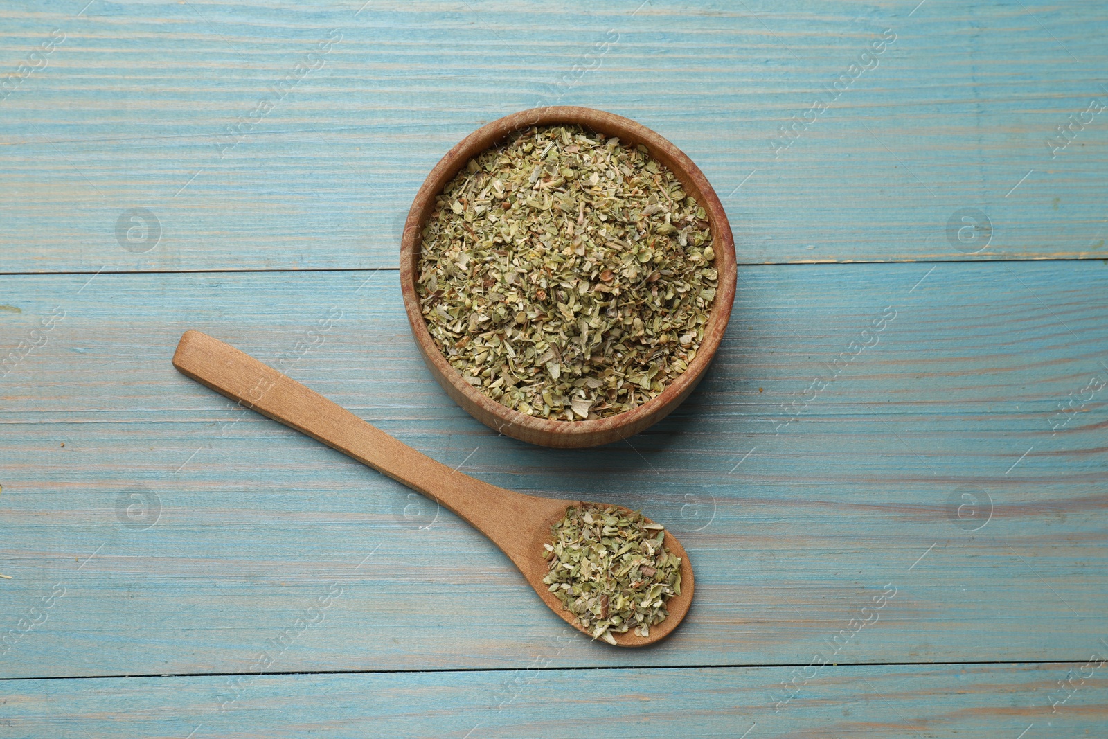 Photo of Dried oregano in bowl and spoon on light blue wooden table, top view