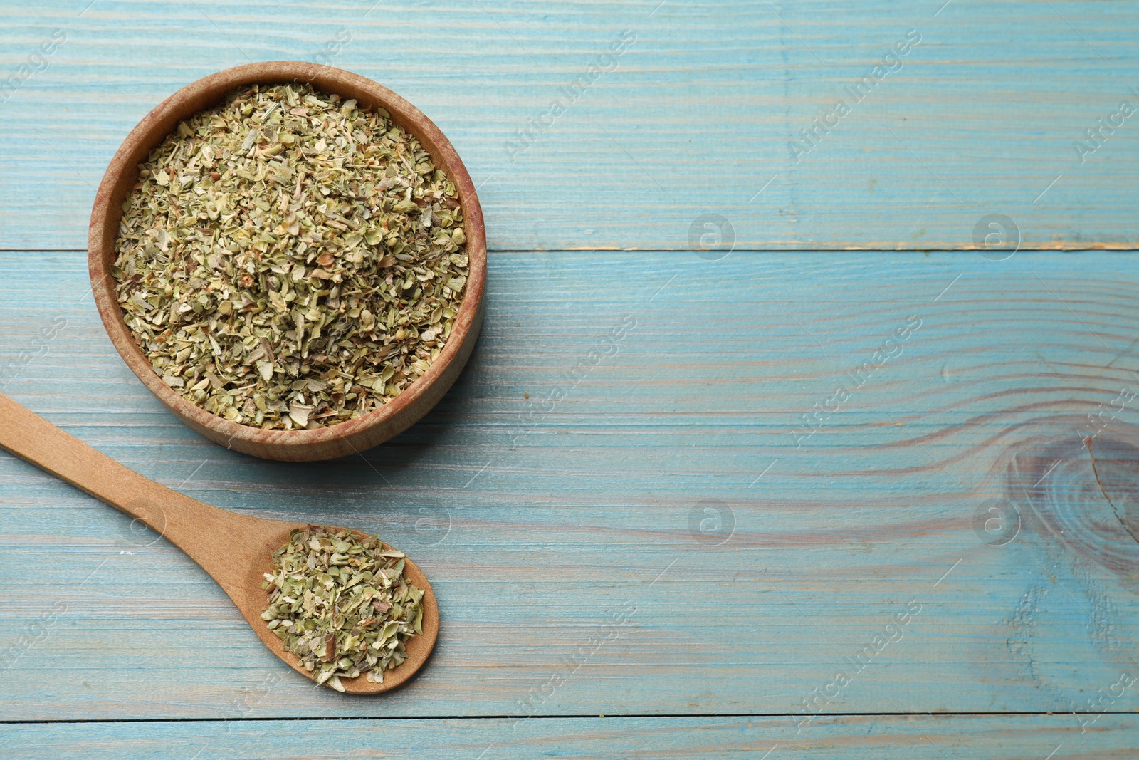 Photo of Dried oregano in bowl and spoon on light blue wooden table, top view. Space for text