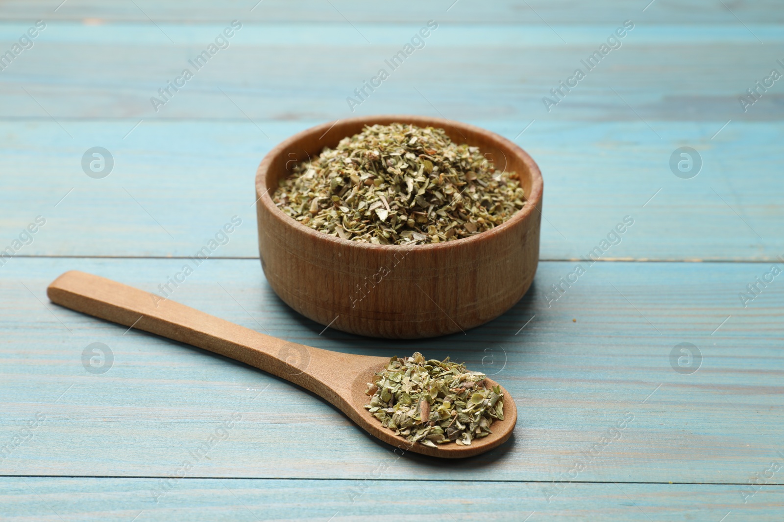 Photo of Dried oregano in bowl and spoon on light blue wooden table, closeup