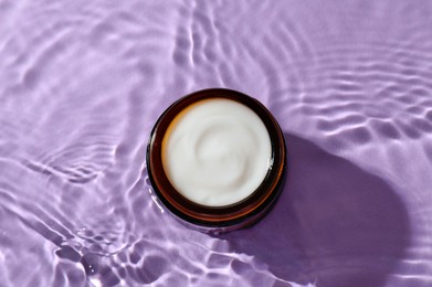 Photo of Jar with face cream in water on violet background, top view