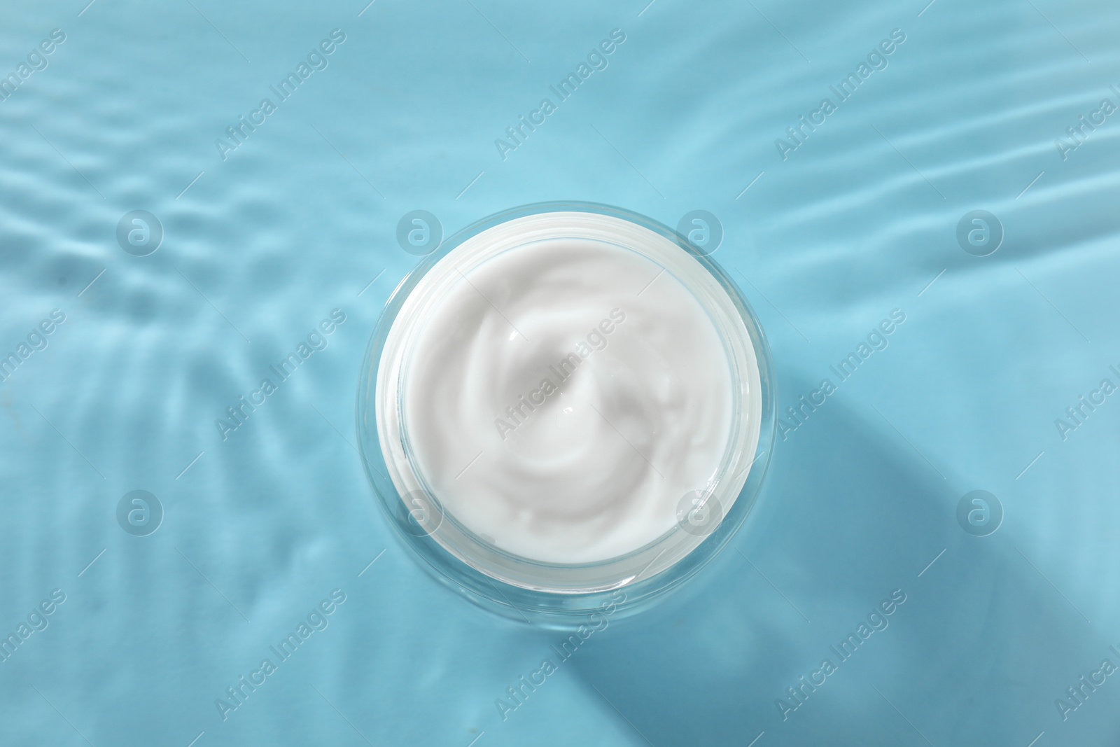 Photo of Jar with face cream in water on light blue background, top view