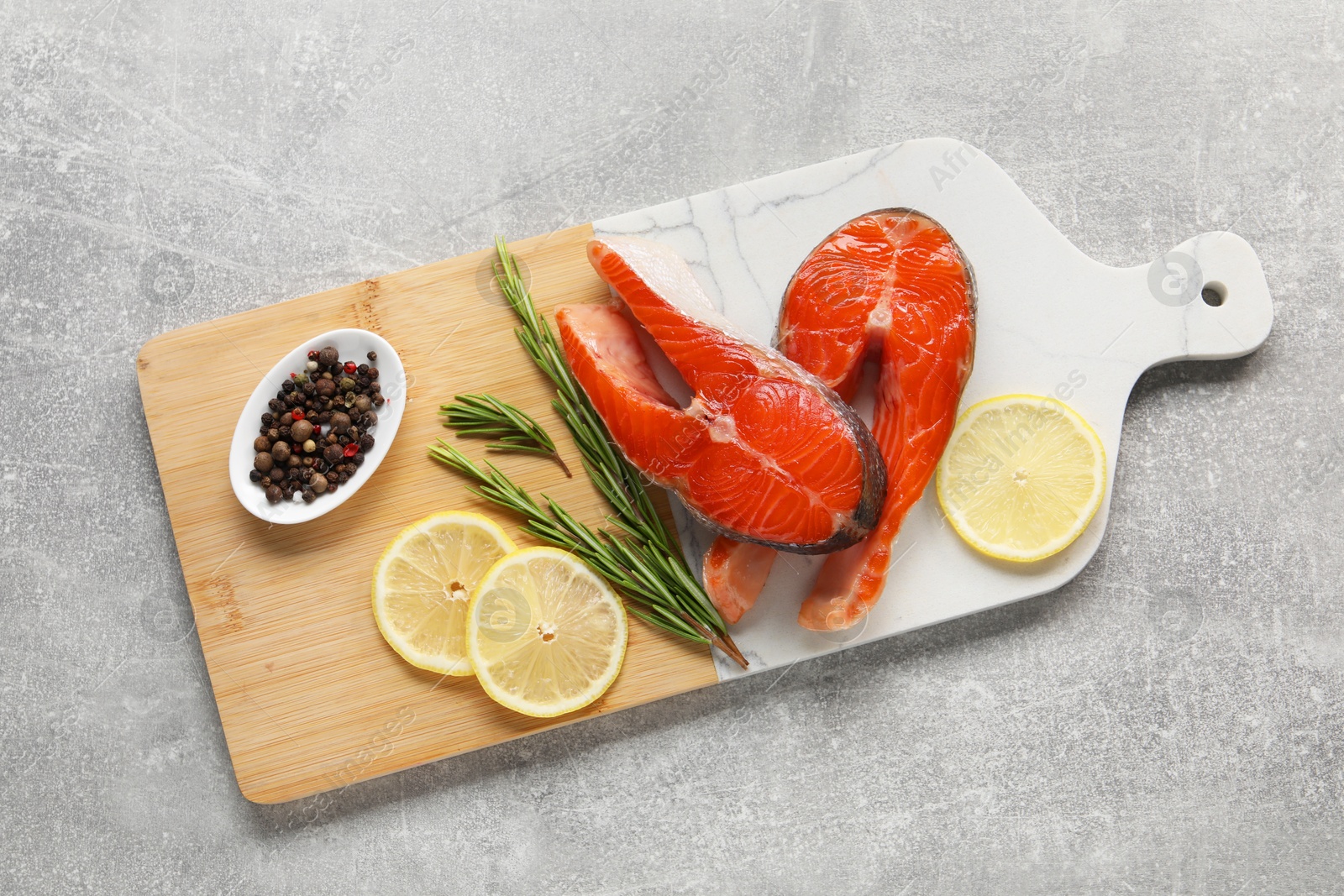 Photo of Board with fresh salmon steaks, peppercorns, rosemary and lemon on grey textured table, top view