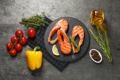 Photo of Tasty grilled salmon steaks, vegetables, oil and spices on gray textured table, flat lay