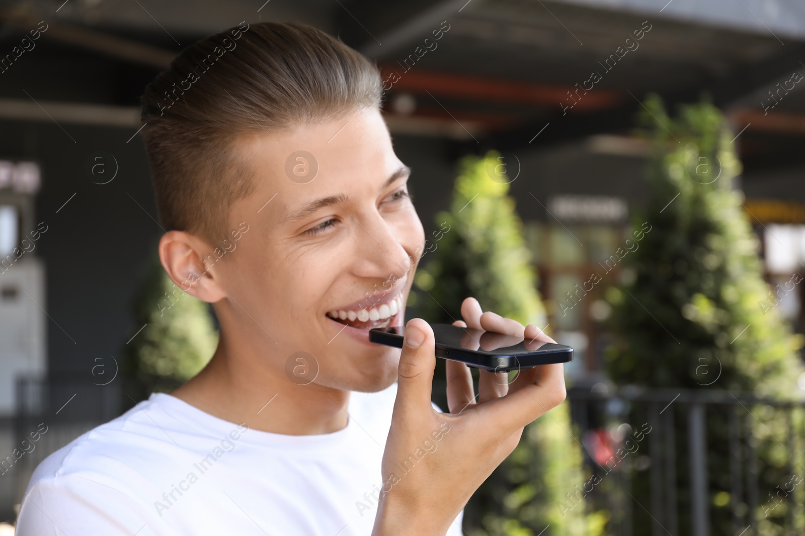 Photo of Young man recording voice message via smartphone outdoors