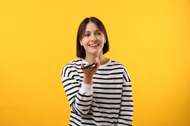 Beautiful woman recording voice message via smartphone on yellow background