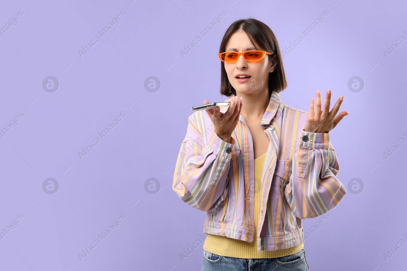 Photo of Beautiful woman in sunglasses recording voice message via smartphone on violet background, space for text