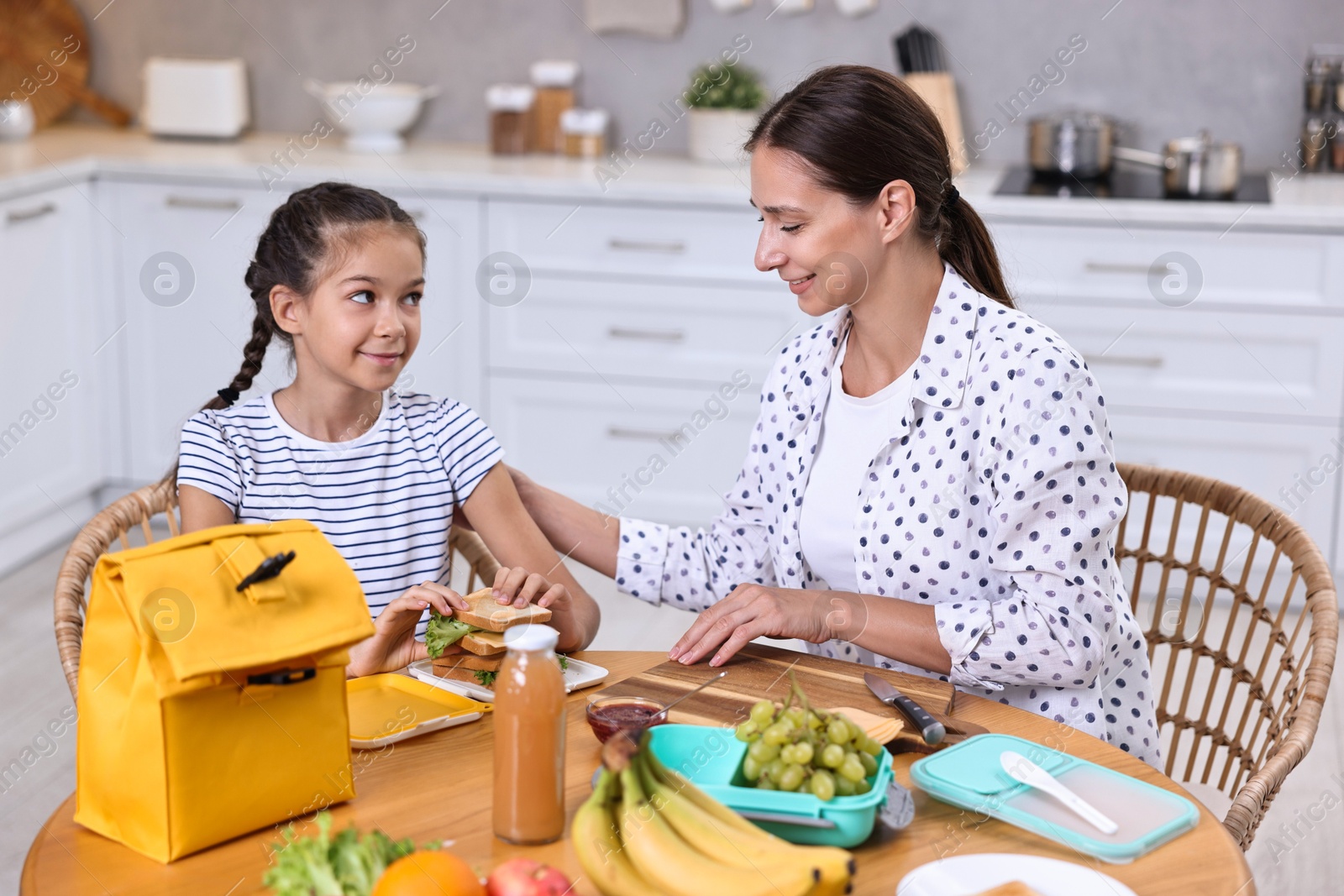 Photo of Cute girl and her mother packing lunch box with healthy products at wooden table in kitchen