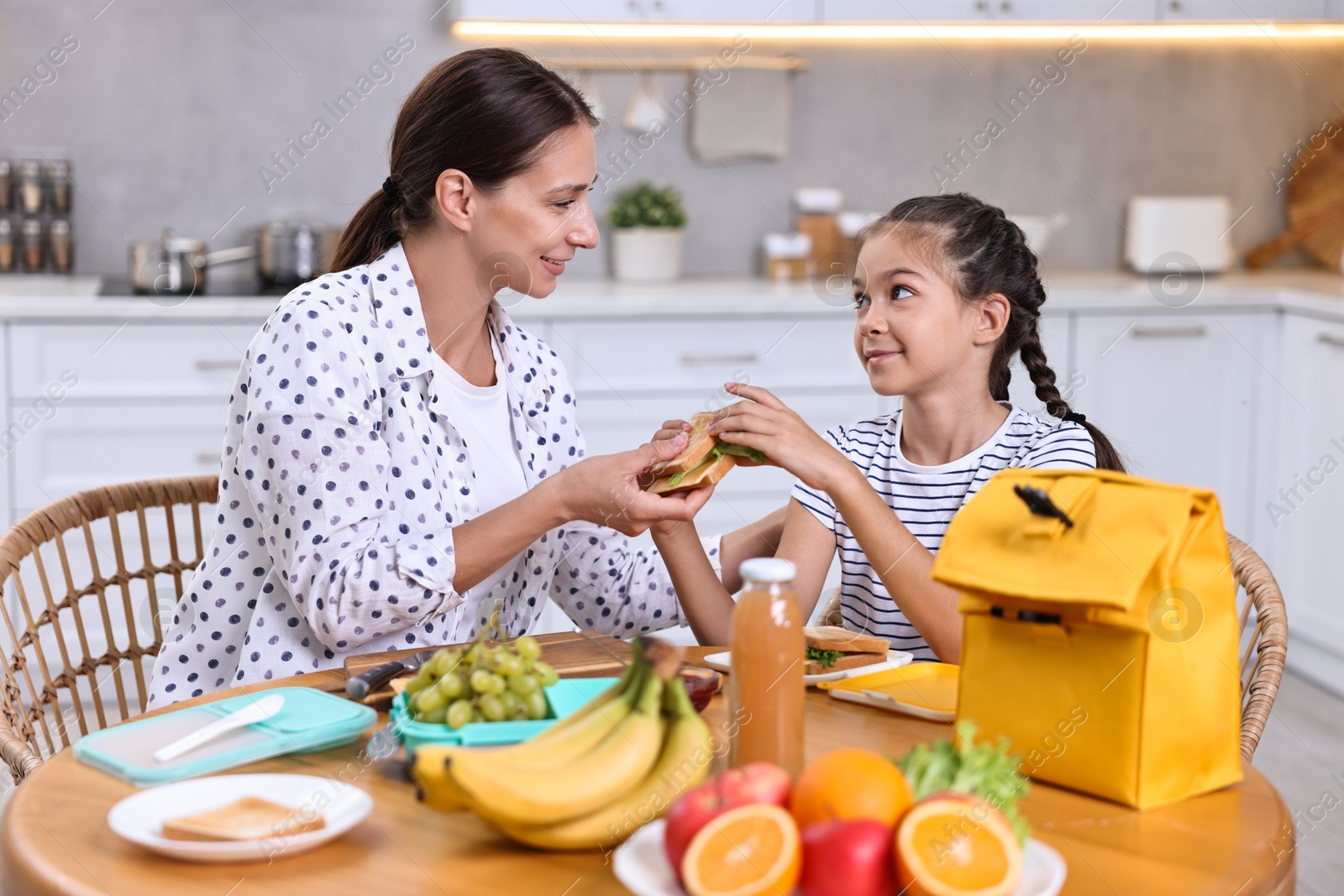 Photo of Cute girl and her mother packing lunch box with healthy products at wooden table in kitchen