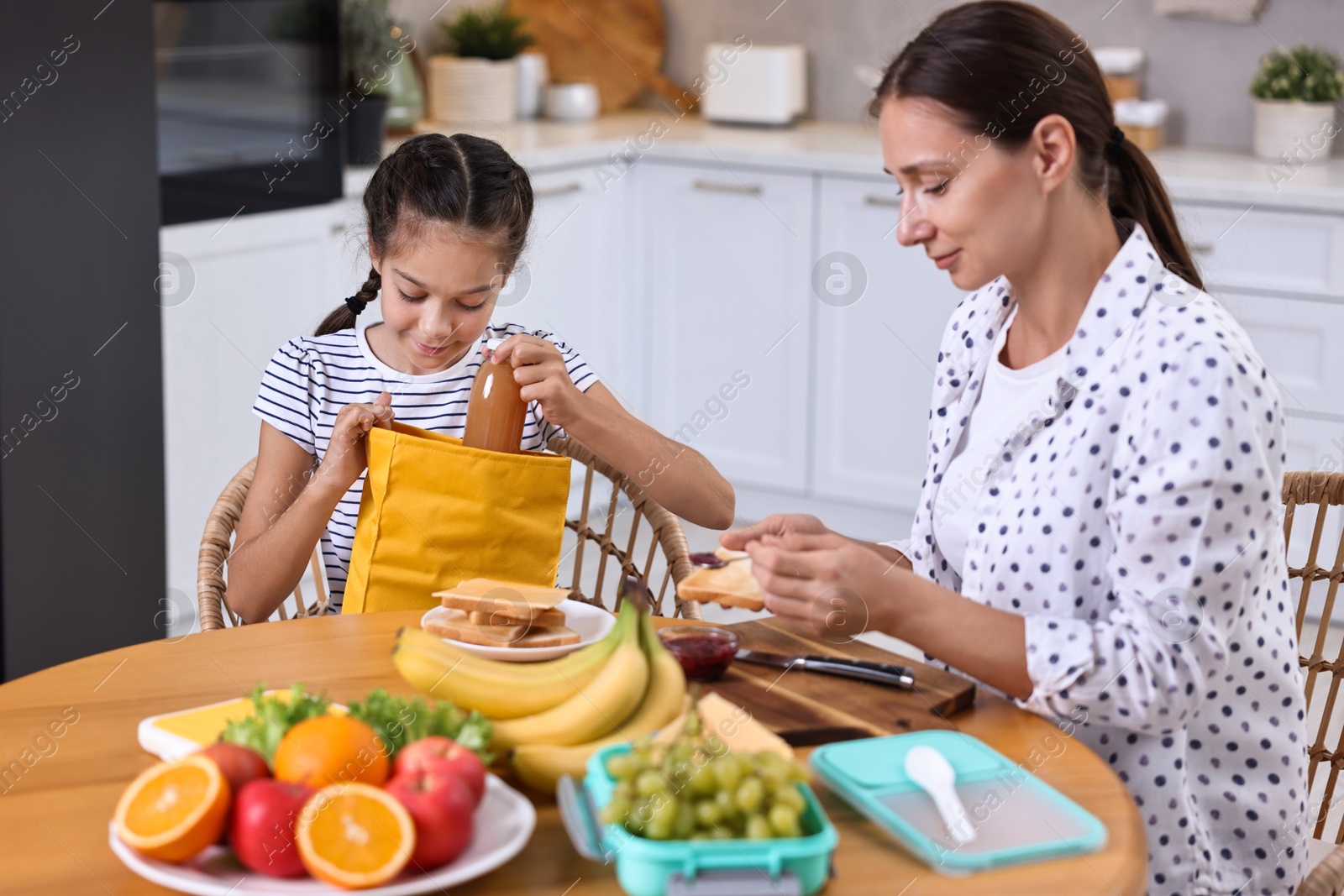 Photo of Cute girl and her mother preparing lunch box with healthy snacks at wooden table in kitchen