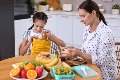 Cute girl and her mother preparing lunch box with healthy snacks at wooden table in kitchen