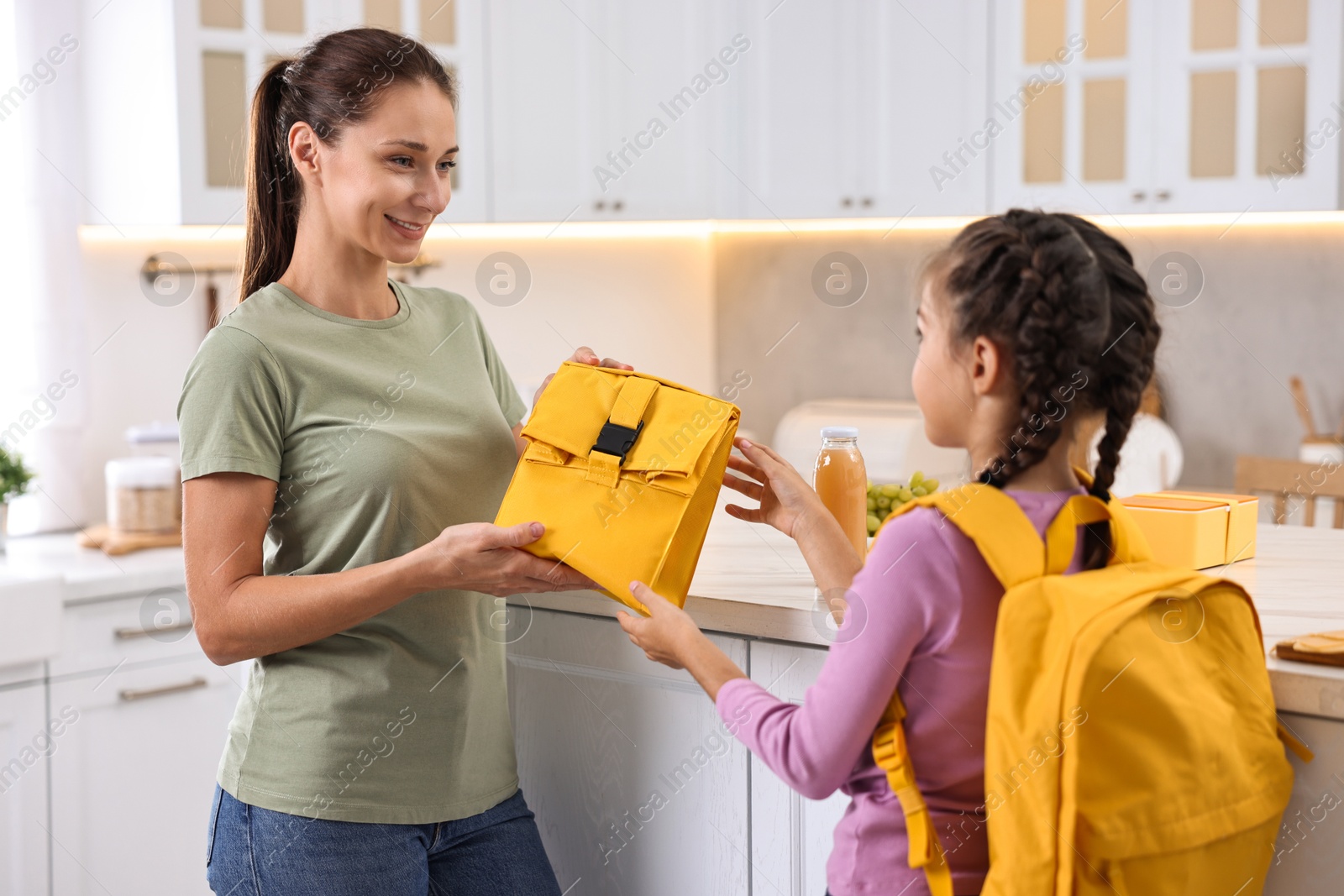 Photo of Smiling mother giving lunch bag to her daughter in kitchen. Preparing for school