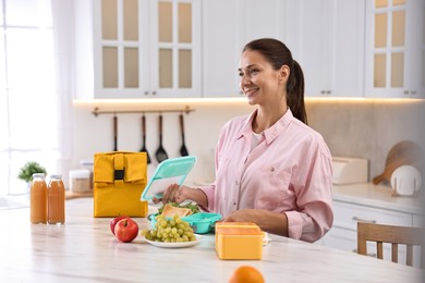 Photo of Smiling woman packing school lunch box with healthy food at white marble table in kitchen