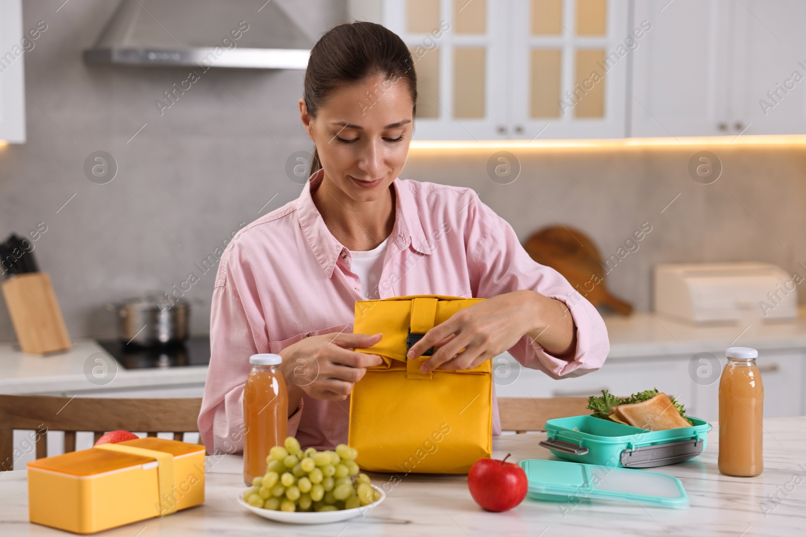 Photo of Woman with bag packing school lunch box and healthy food at white marble table in kitchen