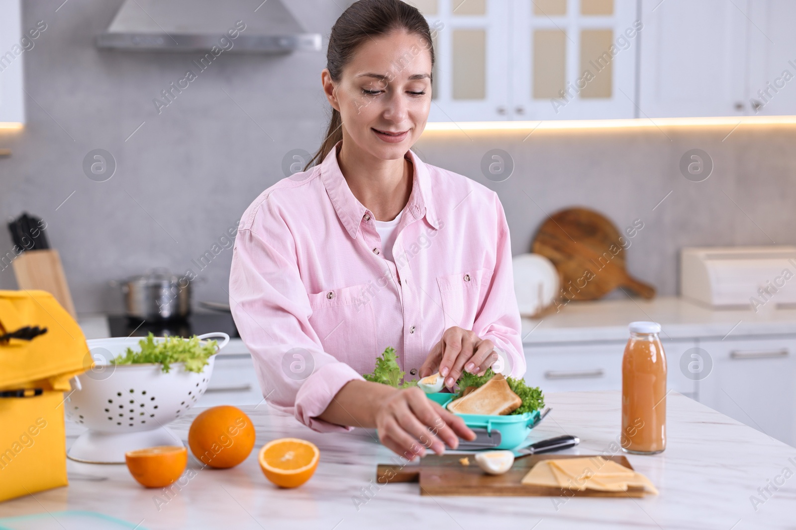 Photo of Smiling woman making snacks for school lunch box at white marble table in kitchen