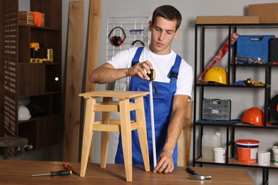 Photo of Man using tape measure while repairing wooden stool indoors