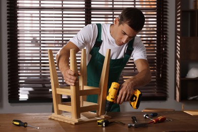 Photo of Man repairing wooden stool with electric screwdriver indoors