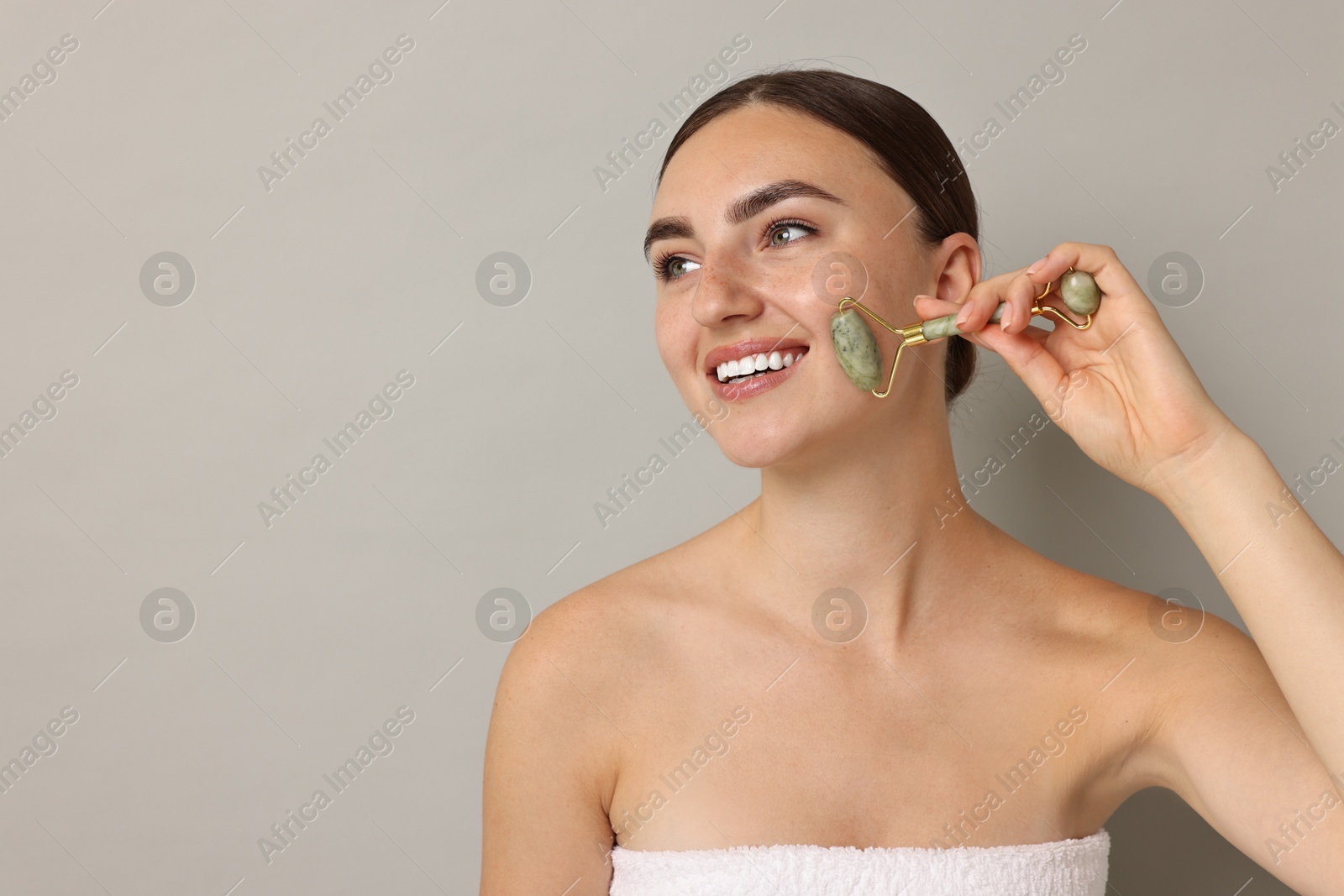 Photo of Beautiful young woman doing facial massage with roller on grey background, space for text