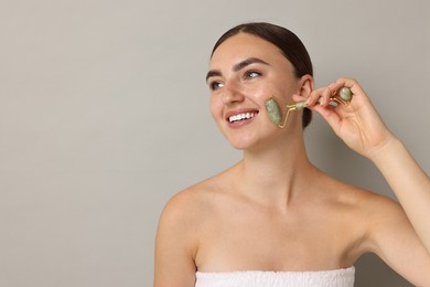 Beautiful young woman doing facial massage with roller on grey background, space for text